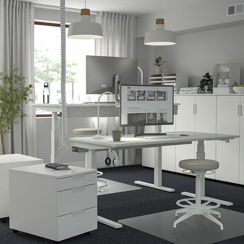 Moderna oficina en casa con escritorio de pie MITTZON, monitores, taburete, gabinete, alfombra, luces, auriculares, planta y ventana grande.