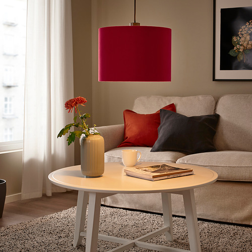 Acogedora sala de estar con lámpara granate sobre sofá beige, almohadas rojas / negras. Mesita blanca con taza amarilla, libro, flor roja.