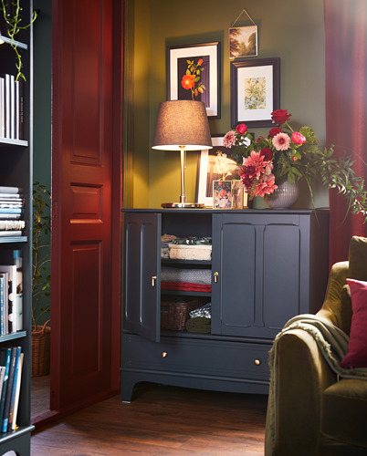 Rincón acogedor con lámpara roja SKOTTORP, gabinete oscuro, jarrón floral, marcos de arte y silla verde.