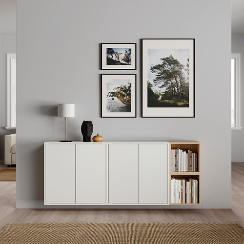 Salón moderno con pared gris y suelo de madera. Armarios EKET blancos, estanterías abiertas con libros, lámpara de mesa y arte de pared con fotos de la naturaleza.