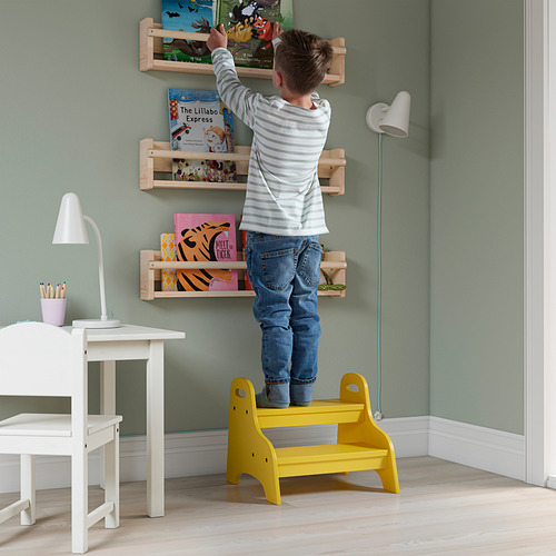 Niño de pie en un taburete amarillo TROGEN, alcanzando libros en los estantes de la pared en una habitación verde.