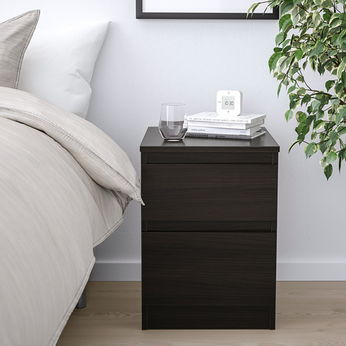 Una mesita de noche negra KULLEN con cajones, un vaso, libros y reloj. Diseño minimalista junto a una cama con ropa de cama vegetal y neutra.