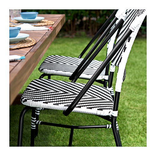 Dos sillas apilables de mimbre blanco y negro se encuentran cerca de una mesa de exterior de madera con platos y vasos.