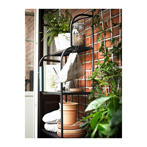 Estantería LÄCKÖ de metal negro con plantas en macetas, libros y un frasco contra la pared de ladrillo.