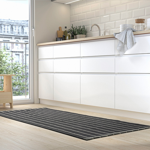 Cocina moderna con alfombra gris RIDSTIG, más pequeña que la puerta, debajo del fregadero. Armarios blancos, toalla a rayas, planta en maceta.