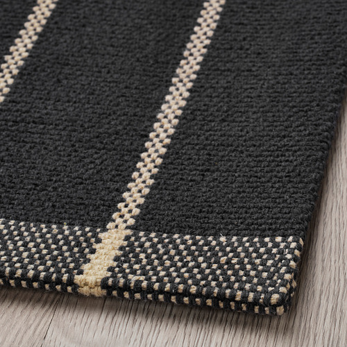 Alfombra de algodón negro texturizado con rayas blanquecinas, colocada sobre suelo de madera. Tejido artesanal de alta calidad visible.