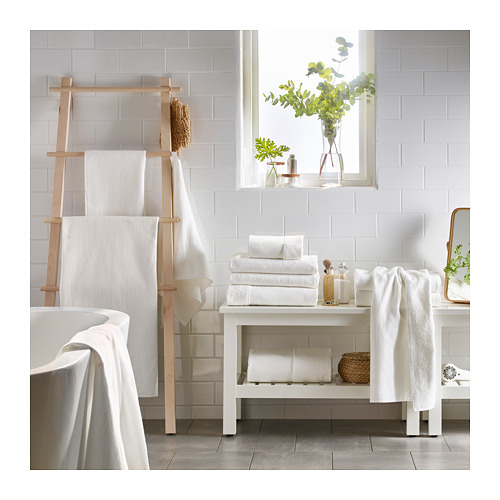 Moderno baño blanco con escalera de toallero de madera, toallas blancas apiladas y plantas en macetas.