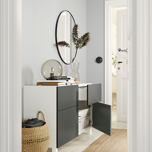 Entrada moderna con mueble de almacenamiento BESTÅ, con cajones blancos y negros, un espejo redondo, plantas y cesta tejida.