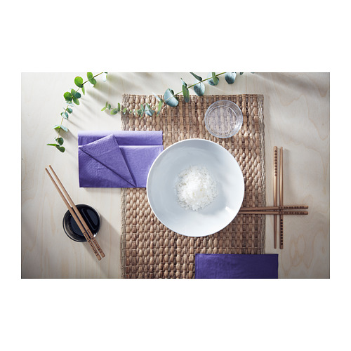 Una mesa de madera con palillos, un cuenco blanco de arroz, servilletas moradas y eucalipto verde. Un mantel individual tejido.