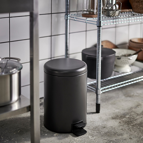 Pequeño cubo de basura de cocina, negro con pedal, junto a una rejilla.