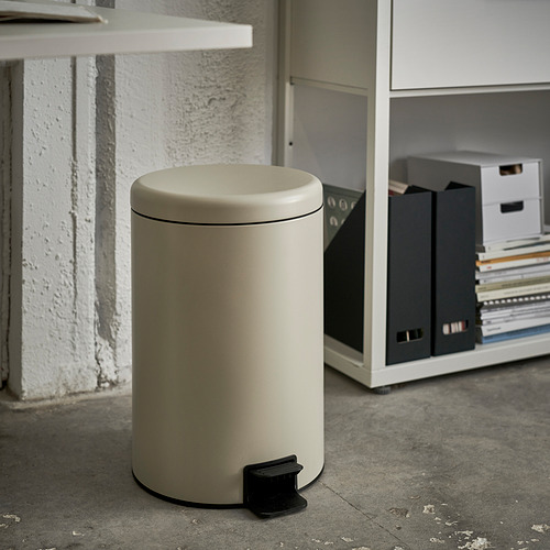 Un cubo de basura cilíndrico de color beige con pedal para el manejo manos libres colocado junto a una pared con estanterías.