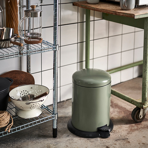 Kitchen scene with a pedal bin on wheels; a shelf with a colander, jar, and cooking tools is beside it.