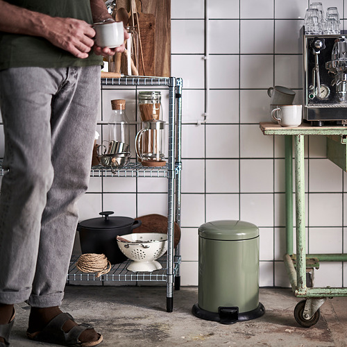 Person holding mug in cozy kitchen with tiled wall, metal shelf, and trash can.