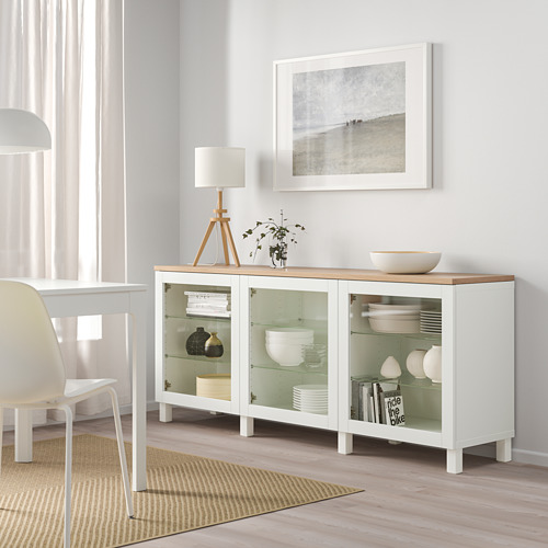 Aparador blanco de ikea BESTÅ con puertas de cristal, que exhibe platos y jarrones, junto a una lámpara, una planta y un cuadro de playa en un comedor luminoso.