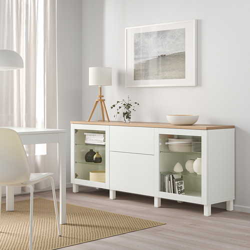 Zona de comedor minimalista con aparador BESTÅ, vajilla, lámpara, planta y foto costera. Habitación llena de luz con suelos y cortinas claras.