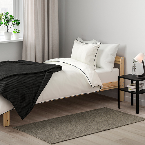 Dormitorio minimalista con decoración en blanco y negro. Pequeña cama de madera con alfombra TIPHEDE, ropa de cama blanca, mesita y lámpara. Cortinas, planta, muro gris.