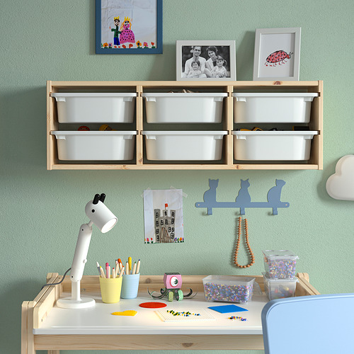 Habitación para niños con almacenamiento de pared TROFAST en madera, con contenedores blancos. El escritorio tiene lápices de colores, decoración y una pequeña lámpara. Los contenedores están en la pared y en el escritorio.