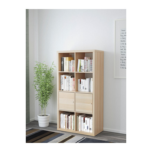 Estantería KALLAX de madera clara, sosteniendo libros, contra una pared. Acompañado de planta alta.