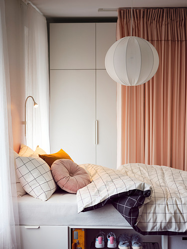 Acogedor dormitorio con ropa de cama de color claro, almohadas de colores, lámpara colgante blanca y caja amarilla debajo.