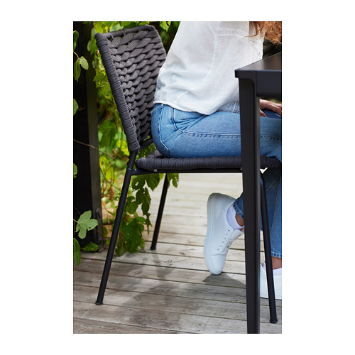 Una persona sentada en una silla de exterior TEGELÖN gris sobre una cubierta de madera, vestida con vaqueros y zapatillas blancas.