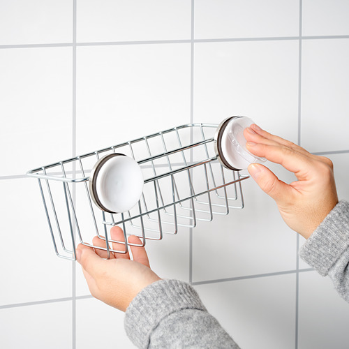 Persona sosteniendo una cesta de ducha KROKFJORDEN plateada con dos jaboneras blancas, contra una pared de azulejos blancos.