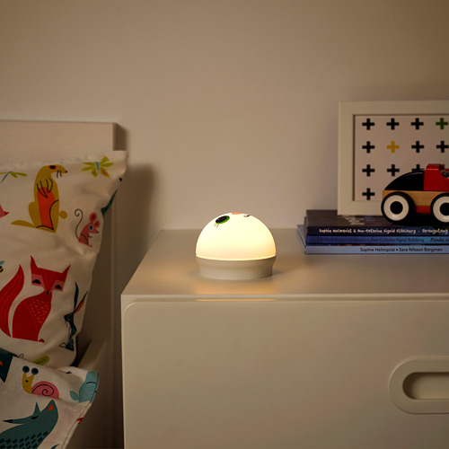 La almohada con estampado Annimal se encuentra en un cajón blanco. Una pequeña luz nocturna redonda llamada KORNSNÖ brilla suavemente junto a libros apilados.
