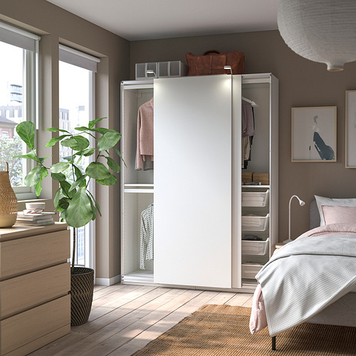 Dormitorio moderno con armario abierto que muestra ropa, estantes y cestas de almacenamiento. Armario PAX en color blanco, suelos de madera, y planta cerca de ventana.