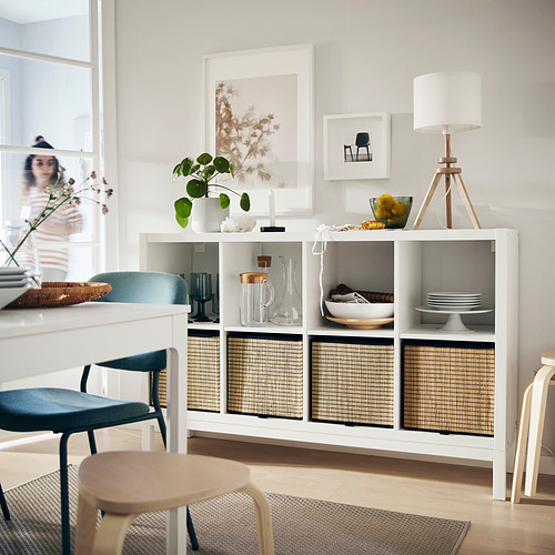Estantería KALLAX blanca con cestas tejidas, elementos decorativos y una lámpara de mesa, colocada contra una pared con arte enmarcado.