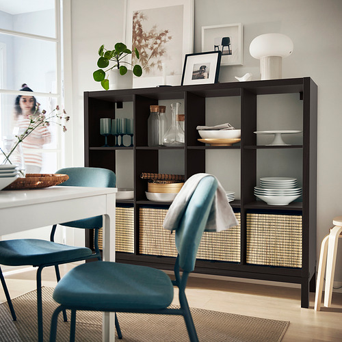 Comedor moderno con sillas verde azulado, mesa blanca y estante oscuro KALLAX con platos, jarrones y plantas.