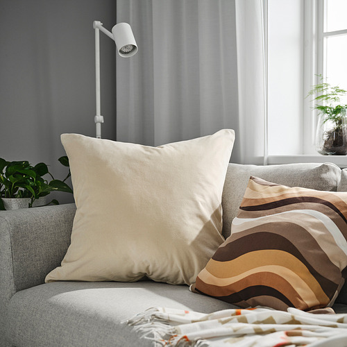 Una gran funda de cojín SANELA beige sobre un sofá gris, con almohadas a rayas y plantas cerca.