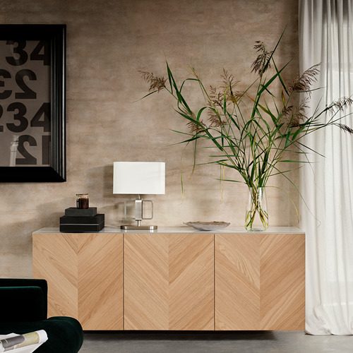 Sala de estar moderna con aparador BESTÅ de madera clara, lámpara blanca, planta alta en jarrón de vidrio transparente, silla verde, marco de fotos negro y cortinas beige.