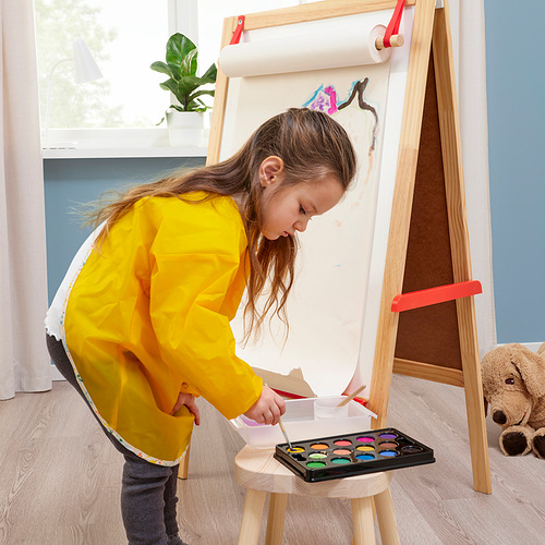 Una niña vestida con un delantal amarillo pinta en un caballete con acuarelas de un kit. Se muestran el caballete y la caja de pintura.