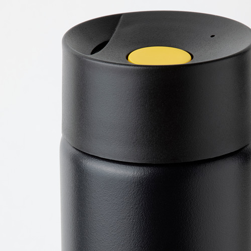 Taza de viaje negra con botón amarillo en la tapa, diseñada para usar con una sola mano, con una superficie antideslizante y un cierre seguro.