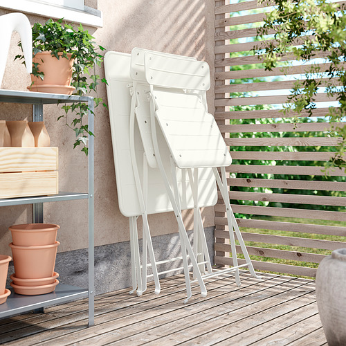 Tres sillas plegables blancas SUNDSÖ apiladas ordenadamente al aire libre sobre una terraza de madera cerca de una pared marrón con estantes que sostienen macetas y plantas de terracota.