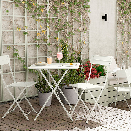 Pequeña mesa de exterior con sillas plegables blancas, vasos verdes y platos en un patio de ladrillos junto a una pared de ladrillos blancos.