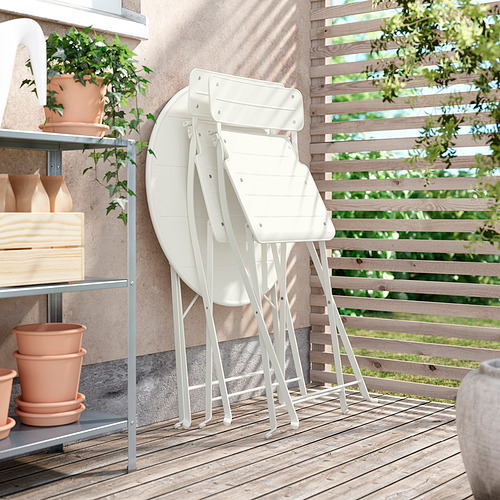 Tres sillas de metal blanco SUNDSÖ apiladas sobre una cubierta de madera junto a una estantería con plantas y macetas.