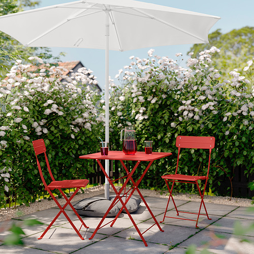 Patio de metal rojo brillante con sombrilla blanca, dos sillas plegables y mesa cuadrada debajo de arbustos en flor, perfecto para relajarse al aire libre.
