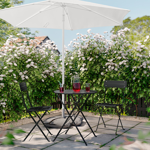 Patio al aire libre con mesa y sillas plegables negras, sombrilla blanca, bebidas rojas, cojines grises, rodeados de flores blancas en flor.