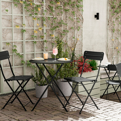 Moderno balcón con mesa y sillas de metal negro SUNDSÖ, rodeado de plantas en macetas.
