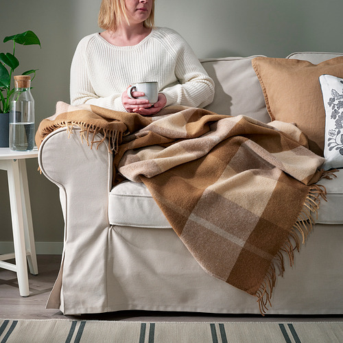 Mujer sentada en el sofá con una manta cálida de lana MYRULL de color marrón claro, sosteniendo una taza, disfrutando de una relajación acogedora.