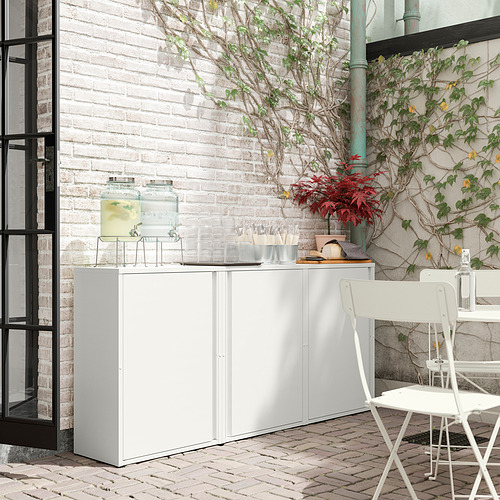 Moderna configuración al aire libre con gabinete SUNDSÖ blanco, pared de ladrillo, plantas verdes y silla blanca.