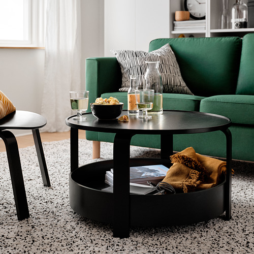 Sala de estar moderna con sofá verde, mesa de centro negra con copas de vino, bocadillos y manta. Alfombra moteada, cortinas blancas.