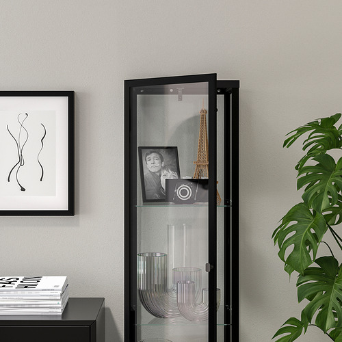 La vitrina con puerta de vidrio negro exhibe artículos, con iluminación integrada y estantes ajustables. A su lado hay una planta de interior.