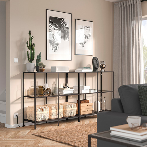 Sala de estar moderna con estante negro VITTSJÖ, plantas, jarrones y libros. Sofá parcialmente visible, ordenado y elegante.