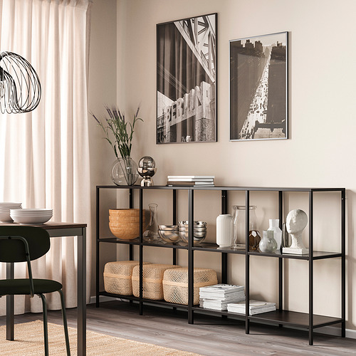 Estantería de cristal VITTSJÖ negro con decoración, jarrones, cuencos, libros; Cerca del comedor, debajo de cuadros enmarcados, cortinas beige.