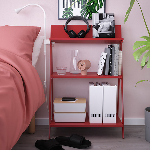 Estante rojo KRÖNIKÖR junto a la cama, con libros, reloj, auriculares, lámpara.