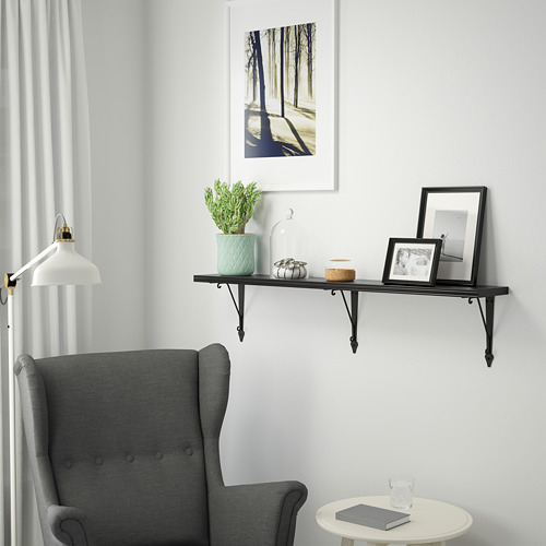 Una acogedora sala de estar con un estante negro BERGSHULT que sostiene una planta, decoración y un marco. Abajo, una mesa sostiene un libro y un vaso.