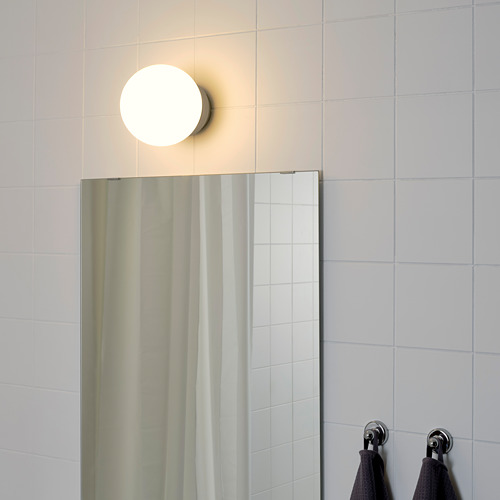 Una lámpara redonda montada en la pared sobre un espejo de baño, que proporciona una luz suave y general. Las toallas negras cuelgan de ganchos de metal a la derecha.