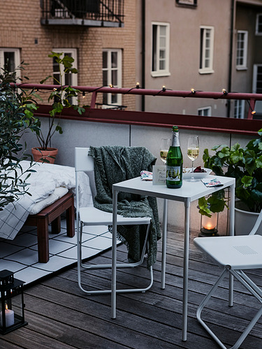 Moderno patio al aire libre con mesa y sillas blancas FEJAN, mantas, vino, plantas y velas, creando un ambiente nocturno acogedor.
