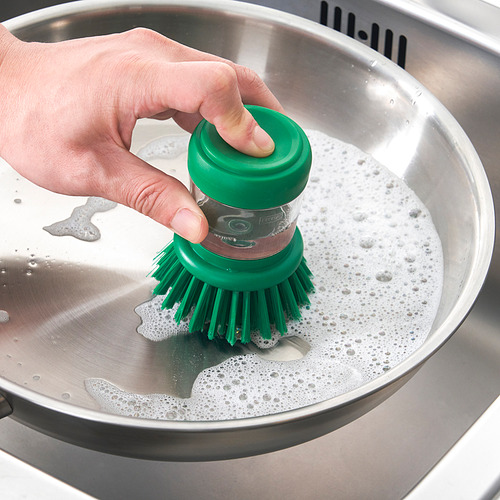 Una mano sostiene un pincel verde sumergiéndolo en un fregadero de acero inoxidable con agua jabonosa, lo que demuestra la capacidad de los cepillos para dispensar jabón.